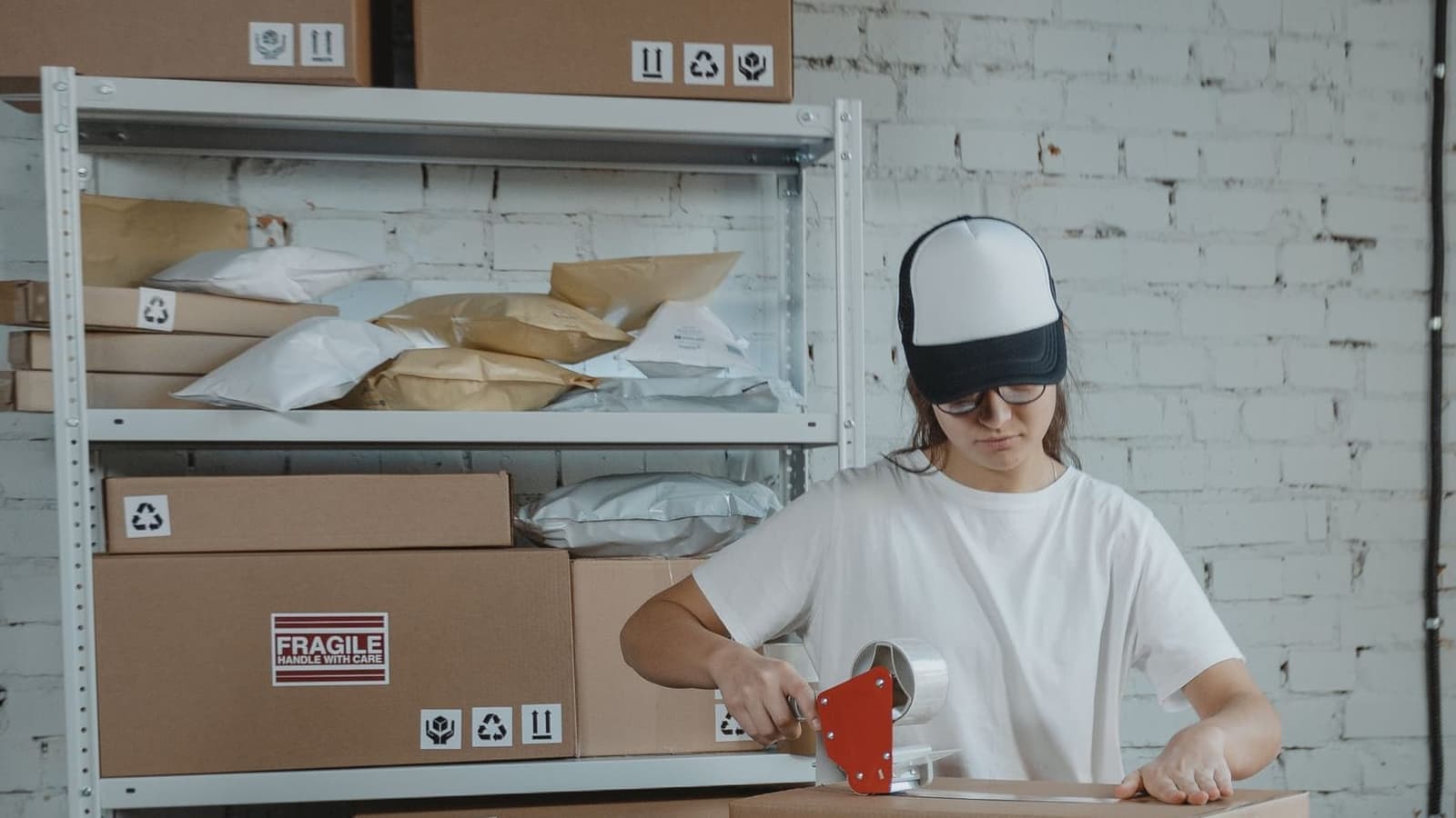 A worker sealing a carton at a packing table, with labeled boxes and stored inventory on the shelves behind the station.