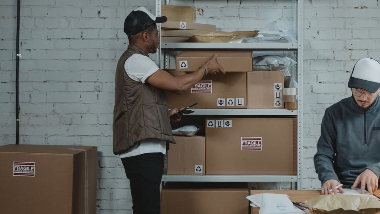 A worker taking a carton from a shelf while another prepares an order at a packing table.