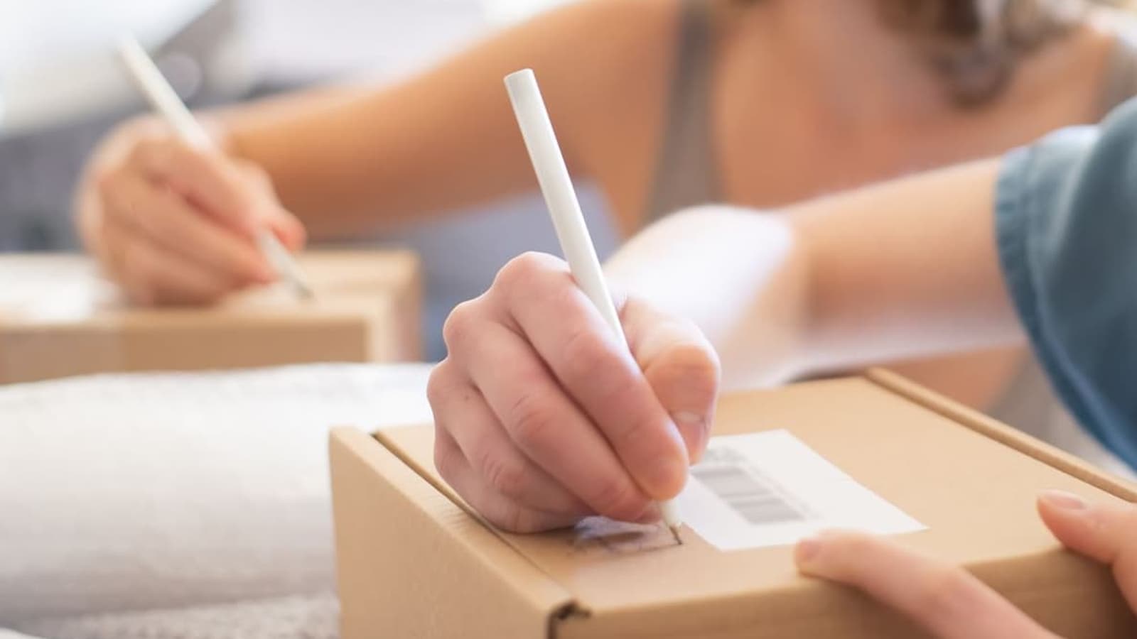 Hands labeling a packed carton, with another person labeling boxes at the same table.