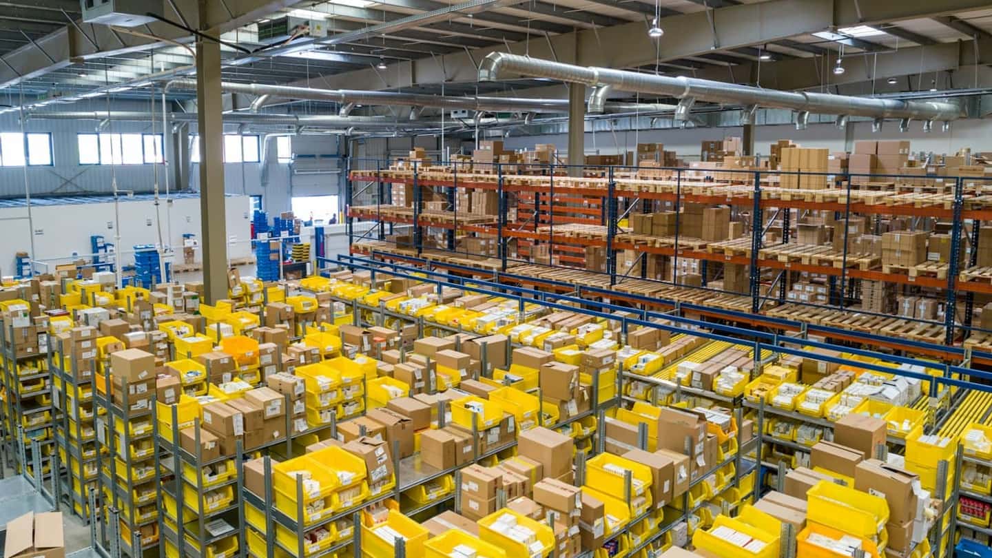 Organized warehouse shelves of cartons and storage bins, with pallet racking behind them.