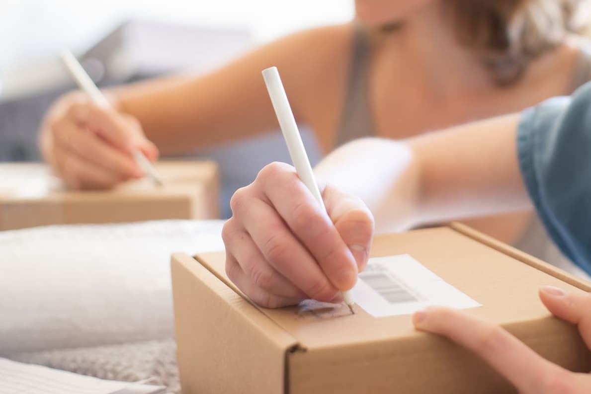 Hands labeling a packed carton, with another person labeling boxes at the same table.