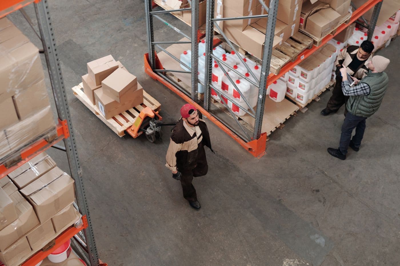 Workers moving a pallet of cartons through a warehouse aisle lined with stored inventory.
