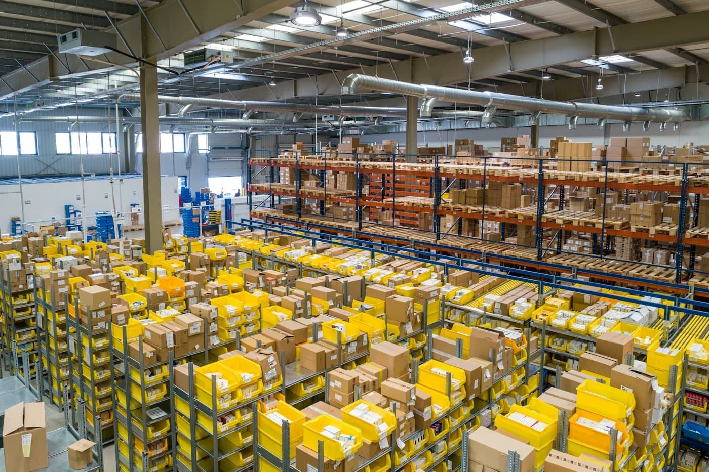 Organized warehouse shelves of cartons and storage bins, with pallet racking behind them.